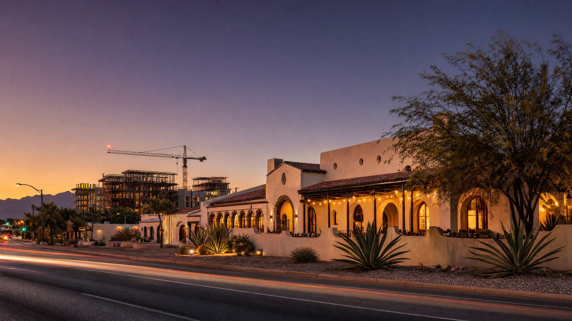 An illustrative dusk scene of a hacienda-style restaurant on a wide desert boulevard — warm lit windows and arched openings under a low tile roof, a covered patio strung with lights, agaves and a mesquite along a low stucco wall, and the steel frames and tower crane of a large construction site rising a few blocks up the street under a violet sky.