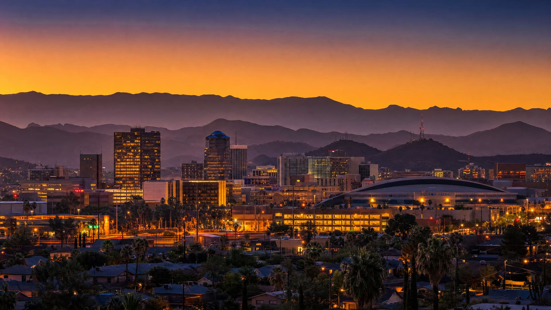 An atmospheric dusk mood image of downtown Tucson — layered mountain ridgelines and a low skyline of blocky building silhouettes, including a shallow arched arena roof, against a deep indigo-to-amber sunset gradient, with warm bokeh lights glowing at street level.