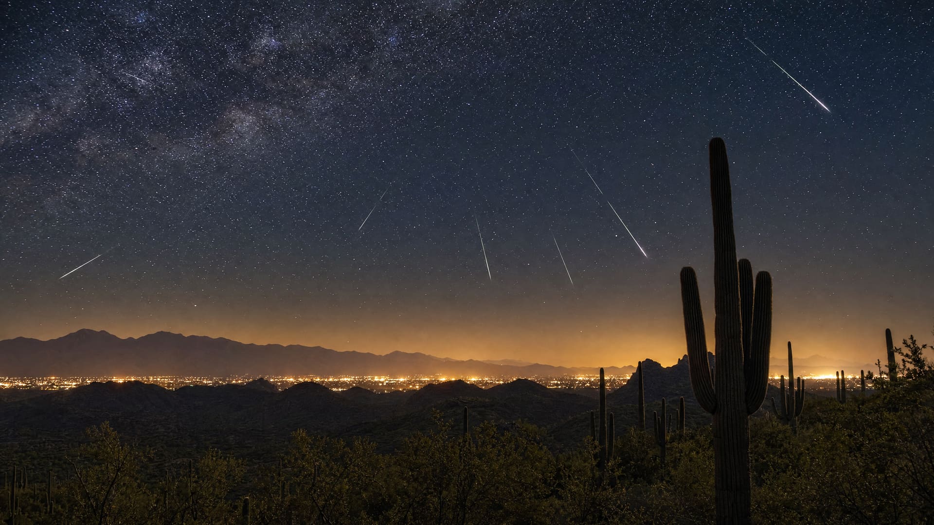 An atmospheric night-sky mood image of the Sonoran Desert outside Tucson — a dark silhouetted mountain ridgeline and saguaro forms beneath a deep indigo-to-black sky scattered with stars and a few meteor streaks, with a faint warm glow along the horizon where the city sits.
