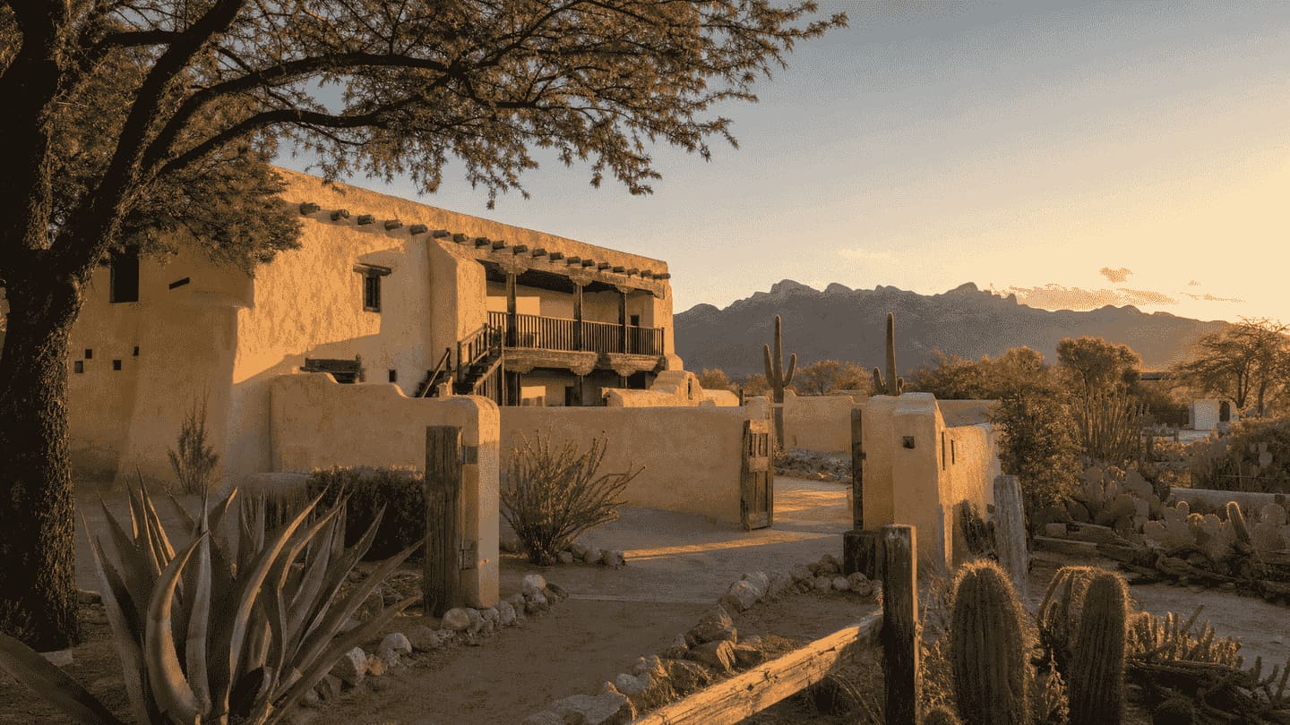 Golden-hour light on a reconstructed adobe presidio compound — a two-story adobe building with a timbered second-floor balcony and projecting roof beams, a low perimeter wall with an open wooden gate onto a dirt path, a mesquite tree overhanging agave and prickly pear in the foreground, saguaros beyond, and a desert mountain ridge on the horizon.